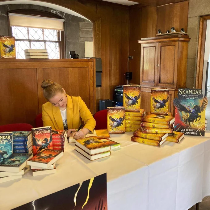 AF Steadman signing copies of yellow chaos trials book with unicorn cover with red and blue Skandar books with epic unicorn covers in shot