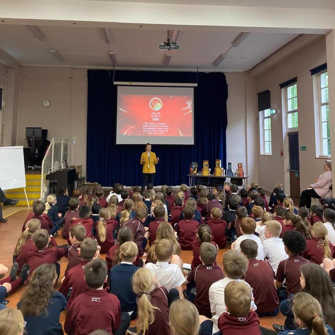 Children in hall listening sitting on the floor while AF Steadman explains about elemental magic - fire symbol in red on the smart board behind her - maroon uniforms