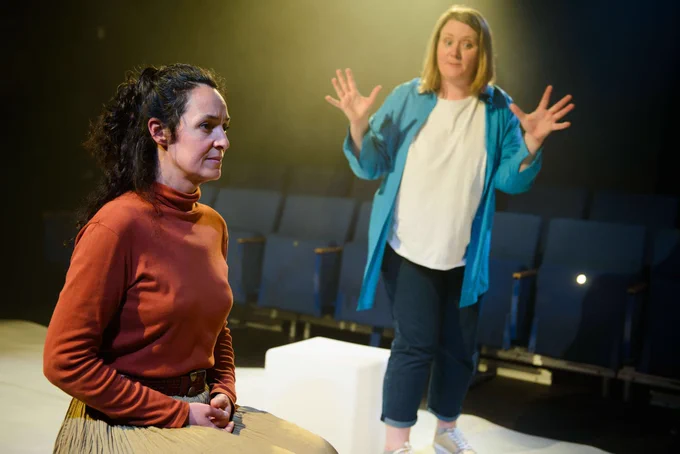 In Omnibus Theatre, Camila is seated on a cube in profile to the camera, while Trine stands, gesturing with her hands as she talks. Camila has long brown hair and is wearing a burnt orange top, while Trine has mid-length blonde hair and is wearing a blue shirt.