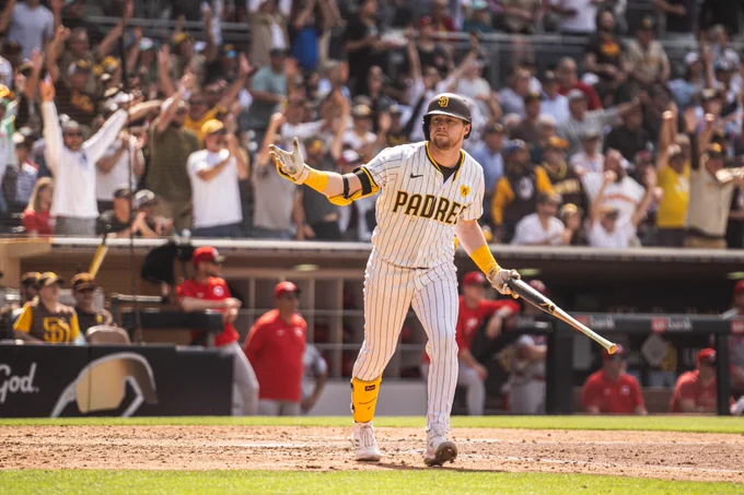 Photo of Jake Cronenworth after hitting a grand slam today. He is looking toward the Padres dugout and shrugging as he gets ready to flip his bat. He is wearing the Padres home white pinstripe uniform. The crowd in the stands behind him is going wild!!!
