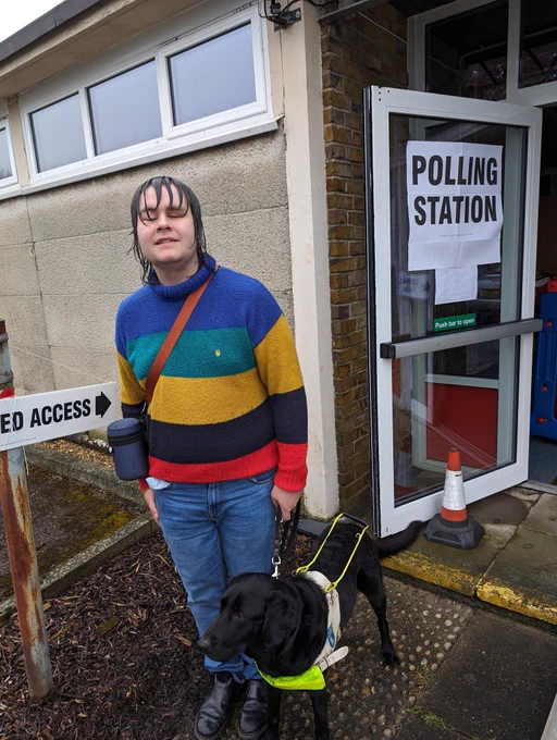 I'm standing outside a building with a sign labeled "POLLING STATION". I am wearing a colorful striped sweater in shades of blue, green, yellow, and red, and blue jeans. I have wet, shoulder-length dark hair and am holding the harness of a black guide dog, Lacey, wearing a high-visibility yellow vest. She is attentively looking up at me. We are standing next to a sign that points to "DISABLED ACCESS". It is a rainy day and the ground is wet.