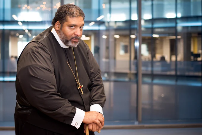 Bishop William J. Barber II, a Black man with a beard wearing his clergy robes, leaning on his cane in front of a window.