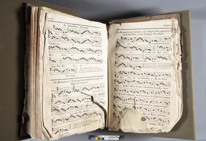 An old book open and showing two pages of musical notes. The pages are torn and pieces missing.