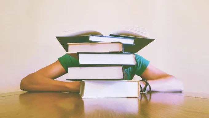 A student hides behind a tower of books on a desk