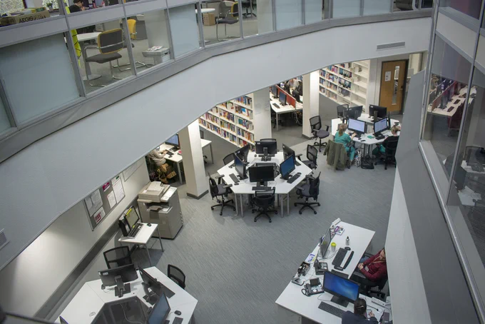 Swindon library from above. PCs are arranged aroun hexagonal tables. Two members of library staff are sat the Help Desk and several students are working on student PCs.