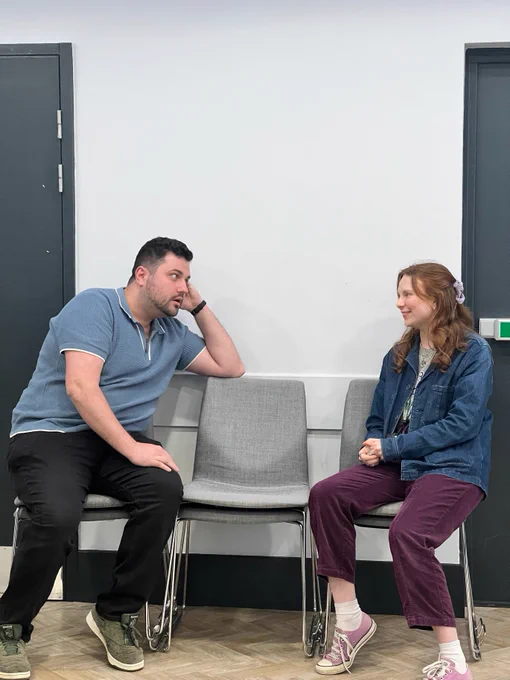 Two people, one in a denim jacket and one in a blue polo top sat on chairs, looking at eachother