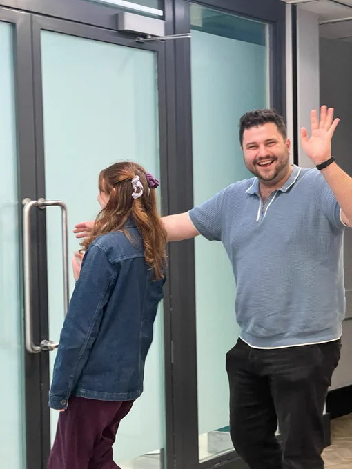 Two people, one in a denim jacke with their back facing the door as if they are leaving and one in a blue polo top waving and smiling at the camera