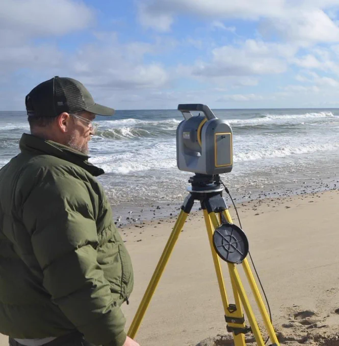 A park scientist stands on a beach next to a LiDAR device on a tripod.