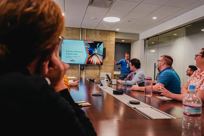 A business presentation in a conference room with a presenter pointing at a screen while attendees look on.