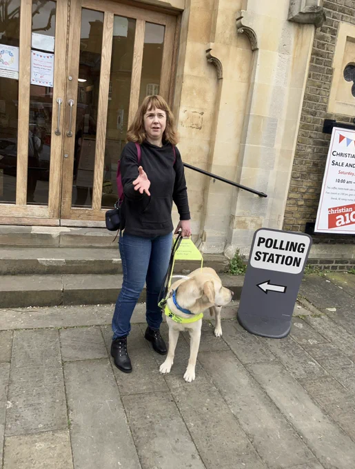 Me a redhead middle-aged woman wearing the black Hoodie and jeans standing holding the harness handle of a large blonde labrador golden retriever dog. In the background is the sign polling station