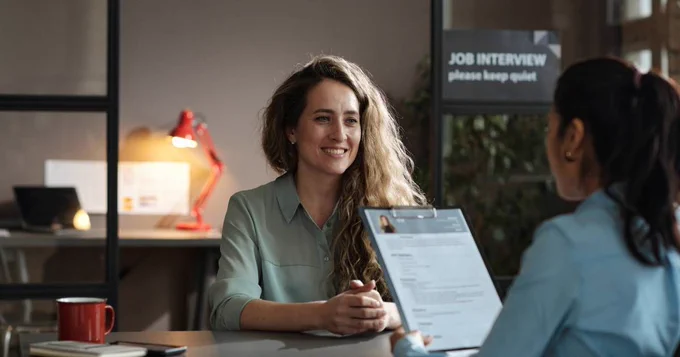 A woman with long hair sitting in an office smiling at an unseen person who is looking at her resume. In the backround is a sign that says “Job interview."