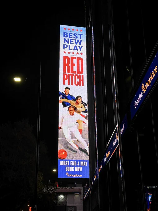 billboard text -
Best New Play
Red Pitch

image -
action shot of three young Black men, one behind the other, all in football poses. the one in the back is wearing a blue tracksuit and jumping to save a shot; the one in the middle is wearing a beige tracksuit with red socks and has just kicked a ball; the one in front is wearing a white tracksuit and is dribbling a red football