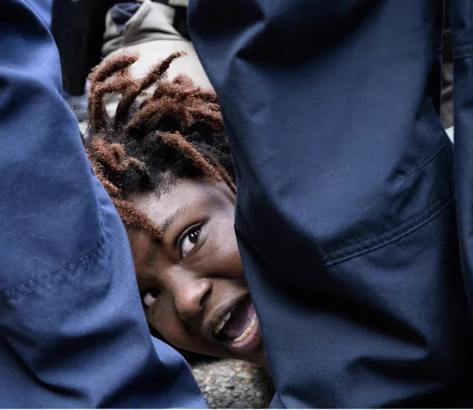 a person with dark skin and dreadlocks pinned on the ground, visible between the blue pant legs of police officers.