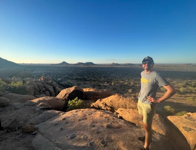 A photo of PLOS ONE Academic Editor Bogdan Cristescu standing on a cliff over looking a vast and sparsely populated expanse of Namibia in the background.  