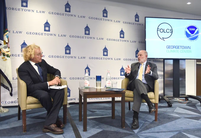 Moderator Thane Rosenbaum and NOAA Administrator Dr. Richard Spinrad sit across from each other in front of a Georgetown Law banner, engaged in conversation. On the television monitor, the logos for Georgetown Climate Center, FOLCS, and NOAA are displayed.