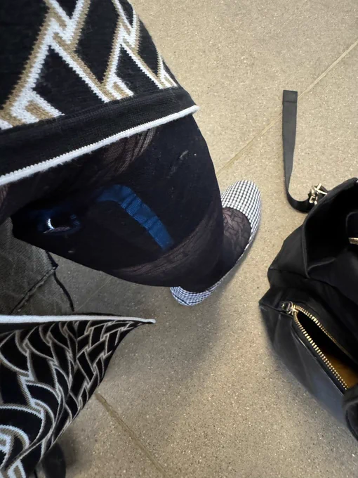 Shiri is showing off her Quell Relief device through a Diane von Furstenberg wrap dress with a black-white-beige basketweave print. The device is worn on her right leg below the knee underneath a geometric-shaped, sheer black pantyhose and a black-and-white checkered ballet flat. On the right edge of the photo, a black bag with gold trim is visible.