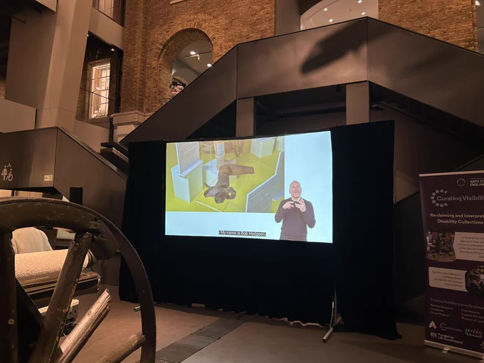 Large screen shows close up image of a wooden artificial arm on display in a museum glass case. There are subtitles and a BSL interpreter