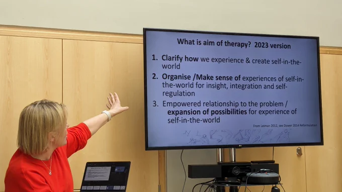 Caroline Greenwood Dower raising her hand towards a screen, talking through slide:

'What is aim of therapy? 2023 version

1. Clarify how we experience & create self-in-the- world

2. Organise/Make sense of experiences of self-in- the-world for insight, integration and self- regulation

3. Empowered relationship to the problem / expansion of possibilities for experience of self-in-the-world'