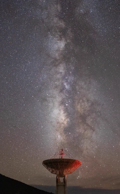 A vertical image features a bright, shimmering portion of the Milky Way galaxy above a massive antenna dish. Thousands of white stars in various sizes dot the sky and vertically down the center is the thick concentration of stars glowing from a soft white to a stronger white, pastel purple, and bits of tan. The dish has a reddish hue, and its antenna is pointed upward with the concentration of stars from the Milky Way almost appearing to flow out of the dish.