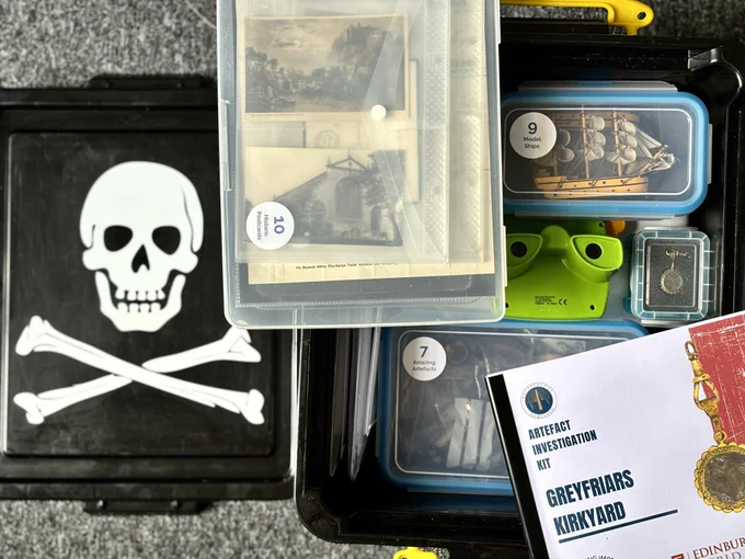 Photo of an opened resource box showing different resources inside - info booklet, viewfinder, archive photos, model ships, mourning jewellery. The box lid with skull and crossbones in it is to the left side.