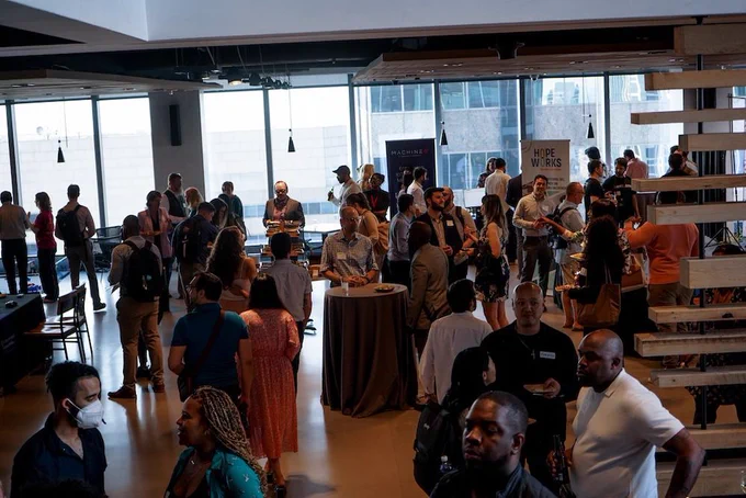 People networking at an indoor event with tables, attendees engaging in conversation, and banners in the background.