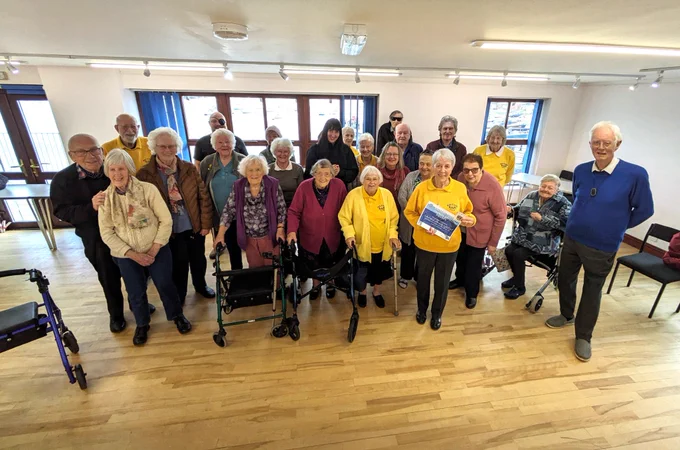 A group of about 30 people stand and smile at the camera. Some of them are wearing yellow jumpers with the words 'Saltash BAPS' on it as a logo. One lady stands in the front holding a certificate.