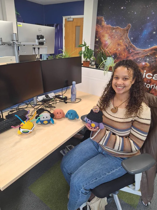 Angelique, a young woman, sitting at her desk and smiling at the camera. Four crocheted mascots are on the desk and she is holding another in her hands. 
