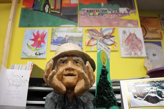 Sculpture of an elderly man with his head in his hands on a desk, surrounded by colorful artwork and a breakfast menu sign.