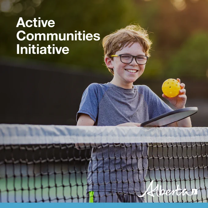 Image shows a boy smiling by the net of a pickleball court holding a paddle and a wiffle ball. The text reads Active Communities Initiative. The white Alberta logo is in the bottom right-hand corner.