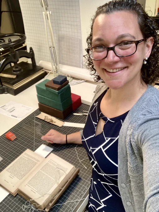Woman with glasses and dark curly hair at a conservation lab work bench with a book being re-sewn. 