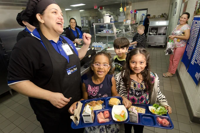 Three kids with lunch trays and a school lunch professional smiling. 