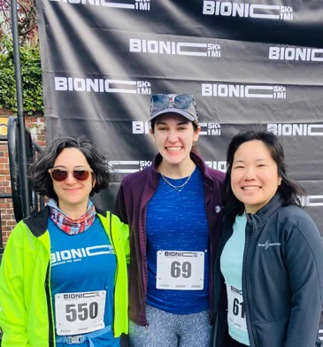 Three women wearing running bibs standing in front of a Bionic 5K step and repeat