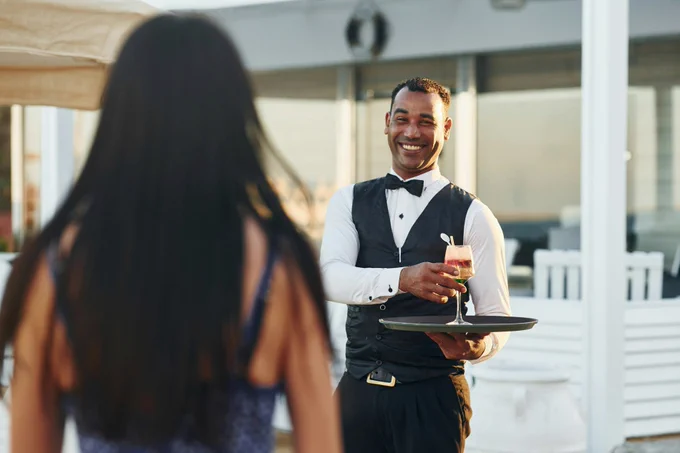 Formally dressed hotel server carrying multi colored drink on tray.

In collaboration with Getty Images
https://unsplash.com/photos/close-up-view-of-drink-black-waiter-in-formal-clothes-is-at-his-work-outdoors-at-sunny-daytime-rFrAzvhM9QI
