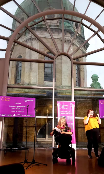 Esther Fox speaking from her wheelchair in front of Curating for Change banners under the dome of the Imperial War Museum London