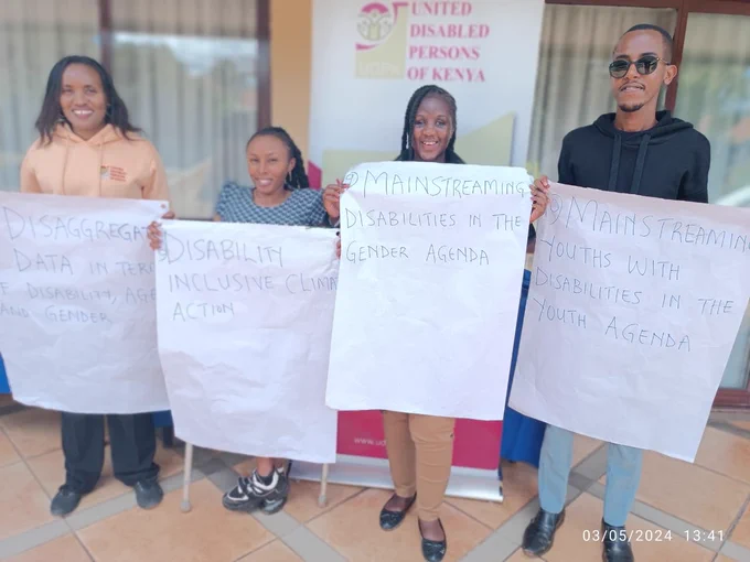Three ladies and one gentleman standing in front of a banner written United Disabled Persons of Kenya.  They are each holding white paper banners with messages on them. The messages include," Disaggregated data,  disability inclusive climate action, mainstreaming disabilities in the gender agenda and mainstreaming youths with disabilities in the youth agenda."