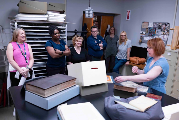 A Notre Dame Preservation employee on the left gestures toward restored books on a work table while library employees in the background watch.