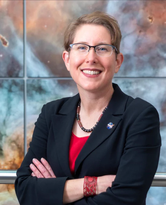 Jane Rigby smiles with her arms crossed in front of a backdrop featuring a Webb image. Jane has short, blond hair and is wearing a black blazer over a red shirt. She has glasses with black rims along the top and is wearing a red bracelet, a red and black necklace, and a NASA meatball logo pin on her lapel. CREDIT: NASA/Jay Friedlander