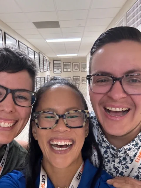 Three workshop presenters standing together posing for a 'selfie' in a school hallway before they present their workshop. All three presenters are smiling and wearing glasses and PHE Canada 2024 Conference lanyards around their necks.