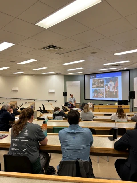 Dr. Lee Airton is presenting at the front of a classroom. Behind them a slide is projected. The slide contains photos of Dr. Lee Airton. Workshop participants are seated behind tables.