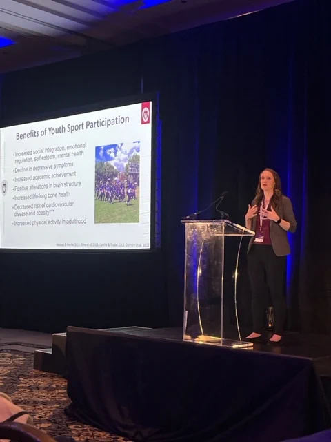 Dr. Julie Stamm standing behind a podium presenting a slide that is titled 'Benefits of Youth Sport Participation' that is projected on a large screen beside them to a ballroom full of International Concussion Summit delegates.