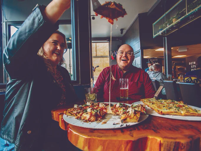 Two people enjoying pizza at Wild Side Brewing in Toledo. The person on the left is pulling a piece of pizza high in the air to show that the cheese separates nice and stringy.