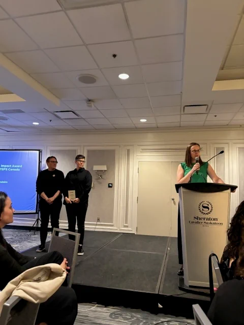 PHE Canada Executive Director & CEO, Melanie Davis presenting the PHE Canada 23-24 Impact Award to Ophea Director of Partnerships and Public Affairs, Tammy Shubat at the 2024 PHE National Conference awards ceremony. PHE Canada President, Jo Sheppard and Tammy Shubat are standing beside the podium Melanie Davis is presenting from.