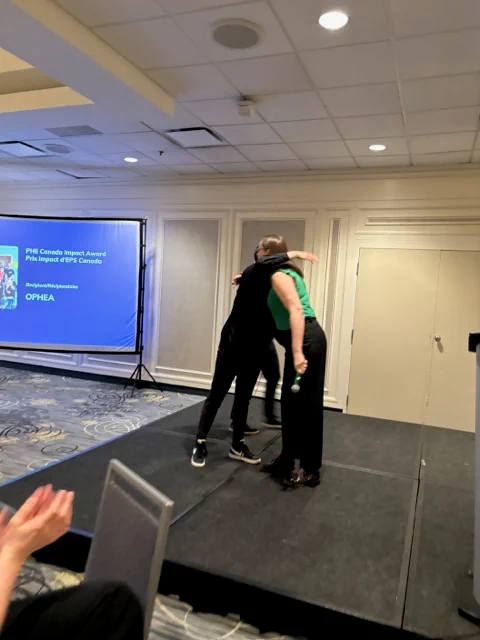 PHE Canada Executive Director & CEO, Melanie Davis presenting the PHE Canada 23-24 Impact Award to Ophea Director of Partnerships and Public Affairs, Tammy Shubat at the 2024 PHE National Conference awards ceremony. Tammy Shubat and Melanie Davis are hugging as Tammy Shubat accepts the award on behalf of Ophea.