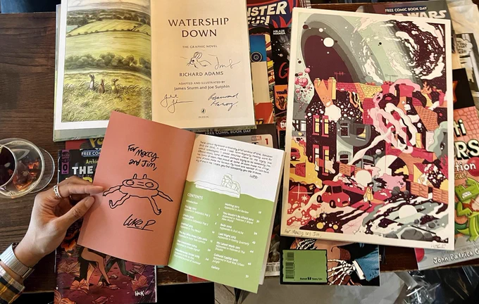 Luke signature and sketch  in his new comic and art print on top of collection of over 10 free comic book day issues on a pub table. Next to Makayla is an Imperial