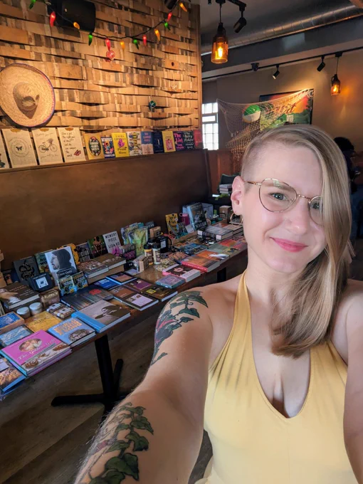 A white woman with blonde hair and gold-rimmed glasses in a yellow dress smiles at the camera, standing in front of a long table laden with books. 