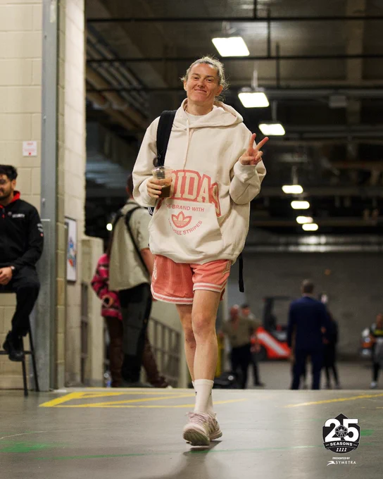 Sami Whitcomb holding up her hand in a peace sign wearing a cream hoodie and salmon shorts, walking through the loading dock at Rogers Place