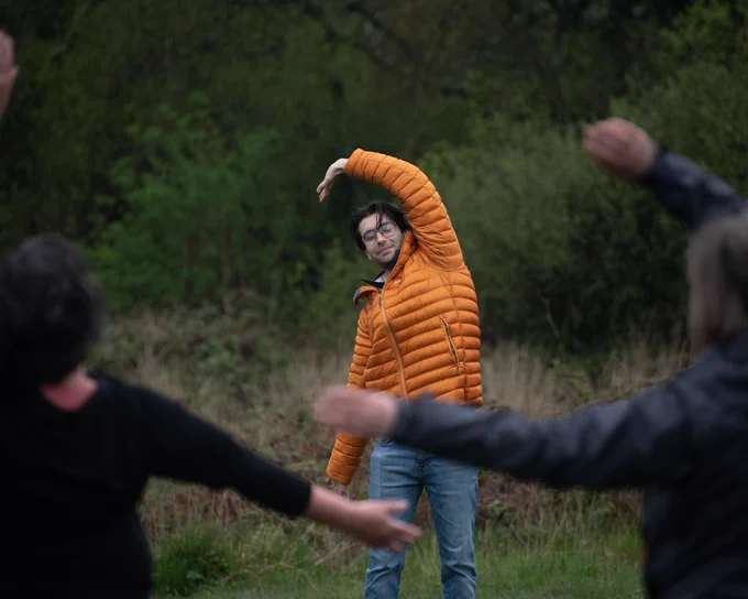 A man in an orange coat doing tai chi in an field surrounded by trees