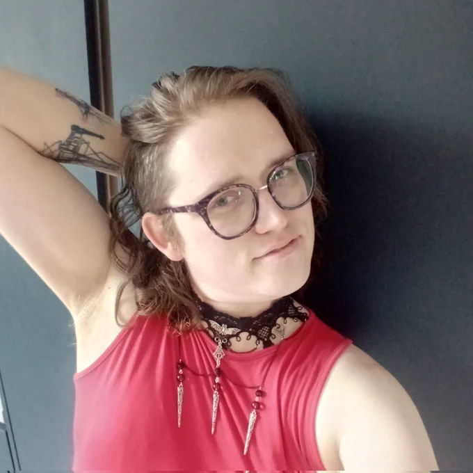 Maddie, a white femme nonbinary person. They're wearing a red dress and an elaborate gothic black choker with metal pendants. She's looking at you seductively. Yes, you specifically.
