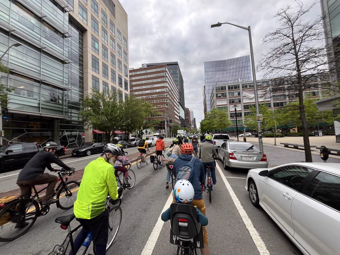 Cyclists of a wide age range (include one child in a carrier) ride across the non-parking width of one side of a city street lined with mid-rise buildings.