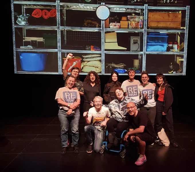 The Touretteshero and Le Grütli team on the stage with a projection of of an image showing a clock with no hands, on a storage rack containing blankets, boxes, tubs, old computers and other items. 